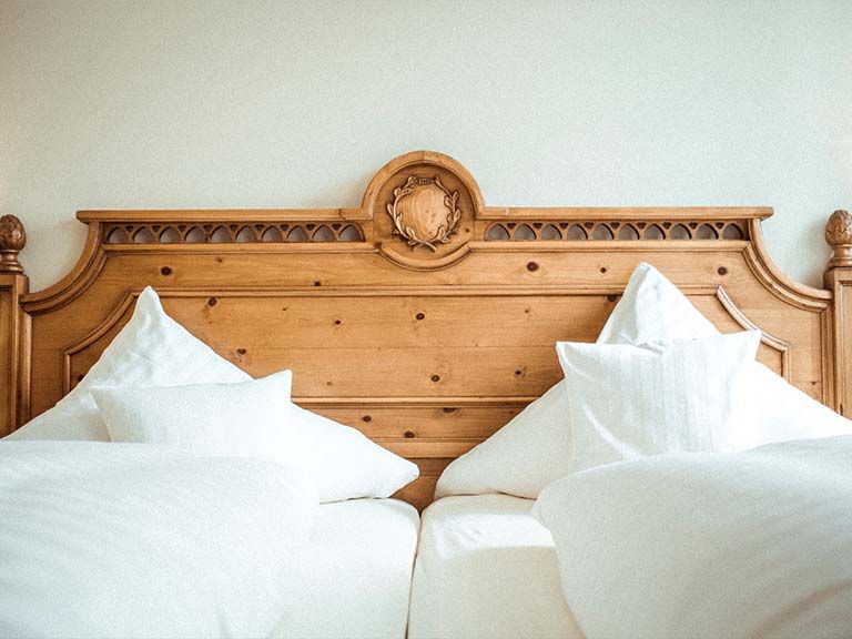 Section of a double bed made of light wood. The headrest is decorated with carved elements. On the bed, fluffy pillows and white duvet covers are scattered.