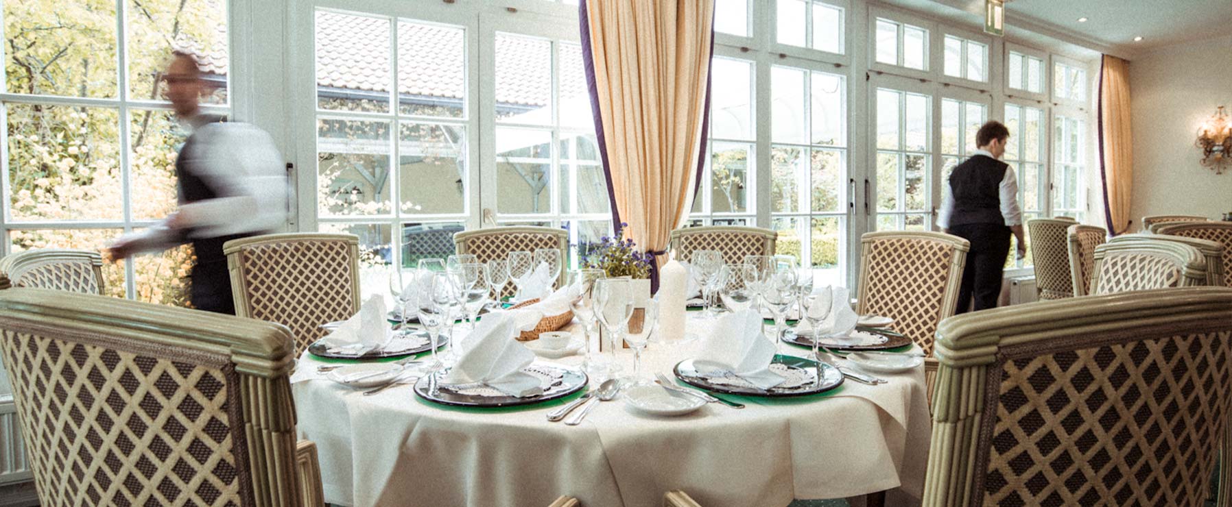 In the foreground, a table set with colorful plates, napkins, and various glasses for water and wine. Waiters are milling around in the background.